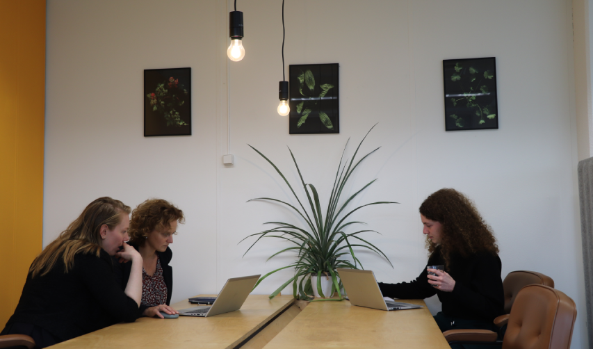 Sfeerfoto van kantoor. Twee personen aan het werk achter de laptop, met daarachter twee personen in overleg.